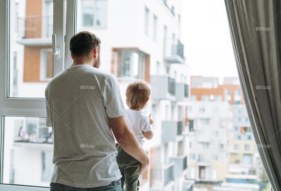 Young man father with baby girl on window sill looking at window at home