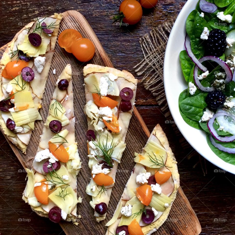 Pizza on tortilla wrap with cashew cheese, olives, cherry tomatoes and fresh dill weed sitting on a wooden cutting board next to a spinach salad.