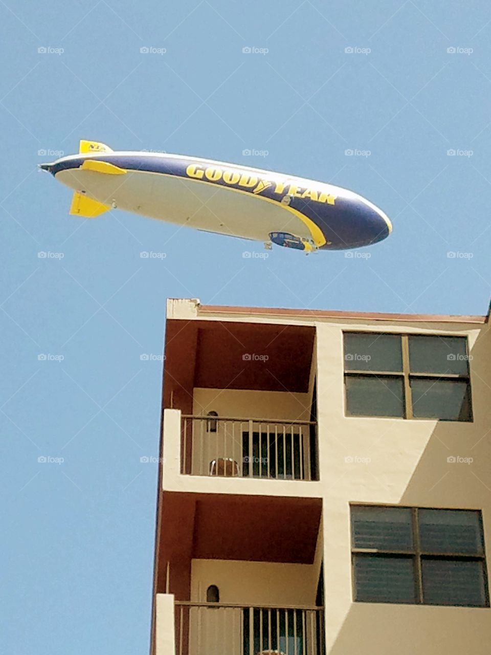 Goodyear Blimp flying low over the corner of a condo, against a clear blue sky. The blimp made no noise as it flew over.