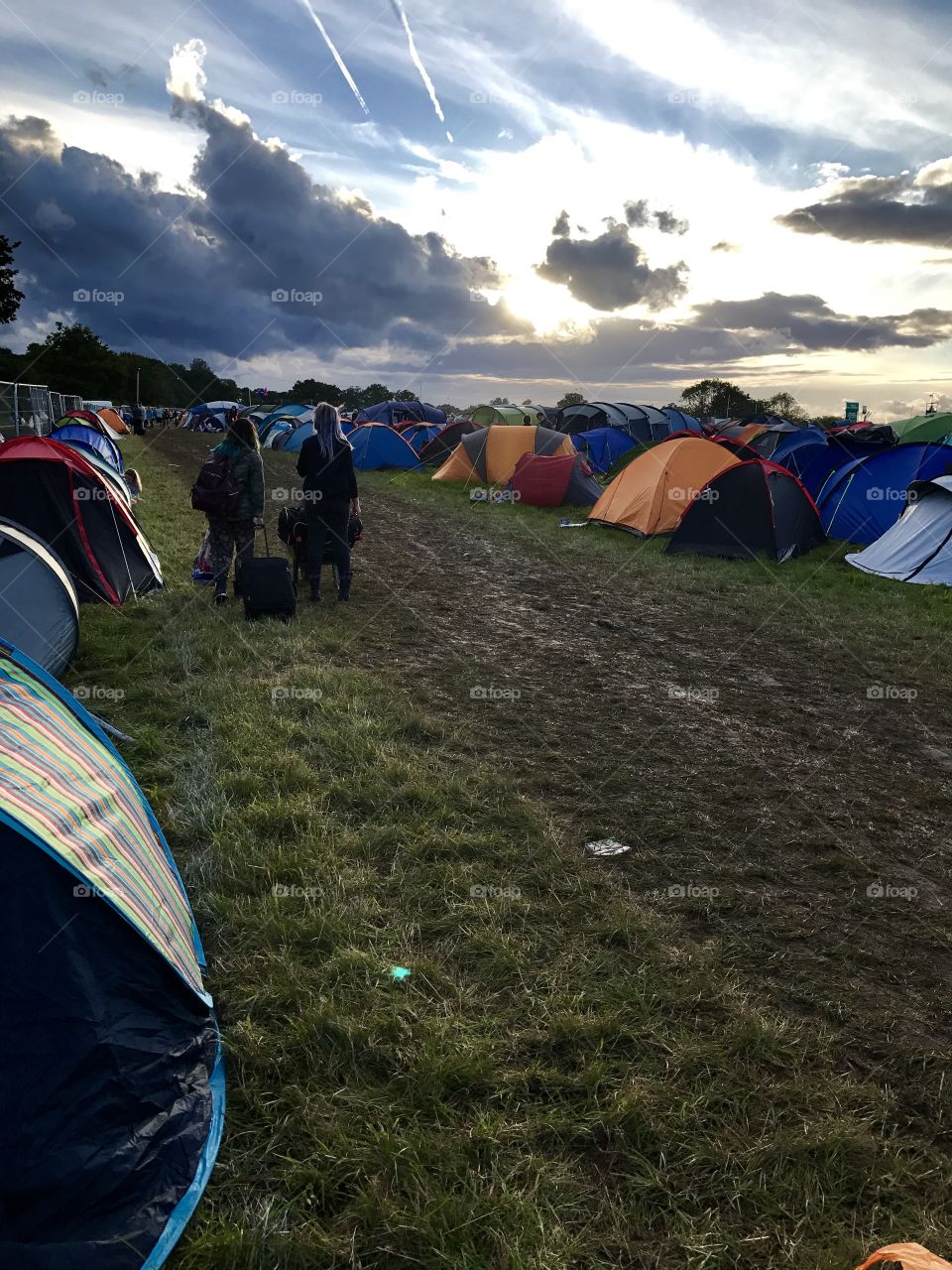 Two girls arrive at a festival and walk through a muddy field surrounded by a mass of other tents with their suitcases as the sun begins to set 