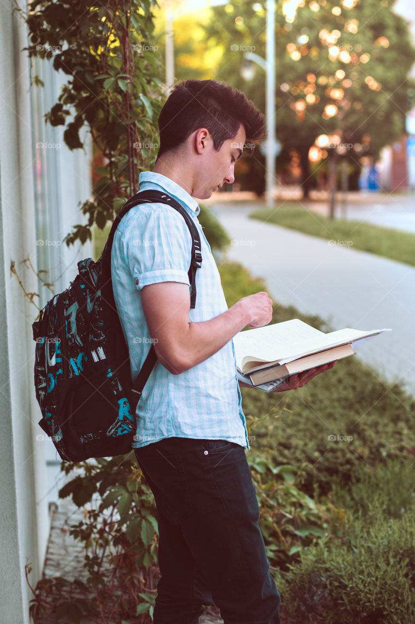 Student holding a notebook and carrying a backpack standing at the front of university building. Young boy wearing blue shirt and dark jeans