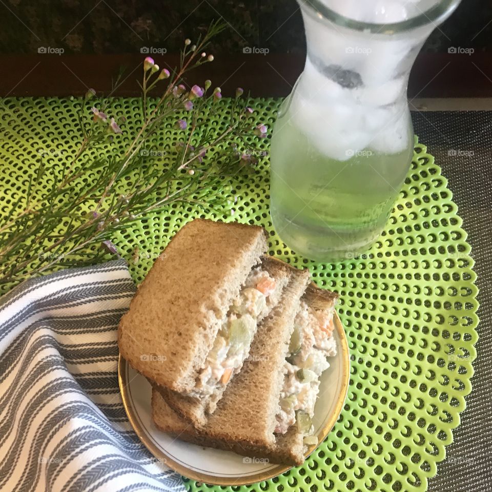 A tunafish salad sandwich with celery, pickles and bell peppers on toasted bread served with a jug of ice water surrounded by lavender flowers on a lime green placemat. USA, America
