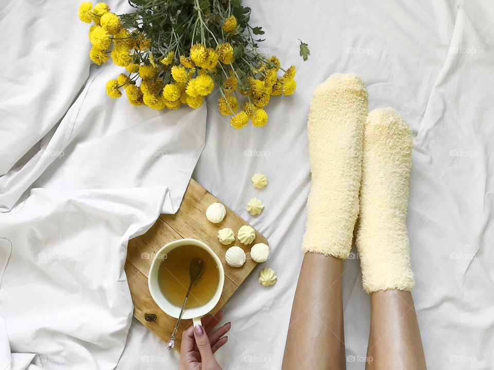 Overhead view of a woman enjoying a tea in yellow cup with yellow candies wearing cozy yellow socks nearby a bouquet of yellow flowers in cozy bed with white sheets 