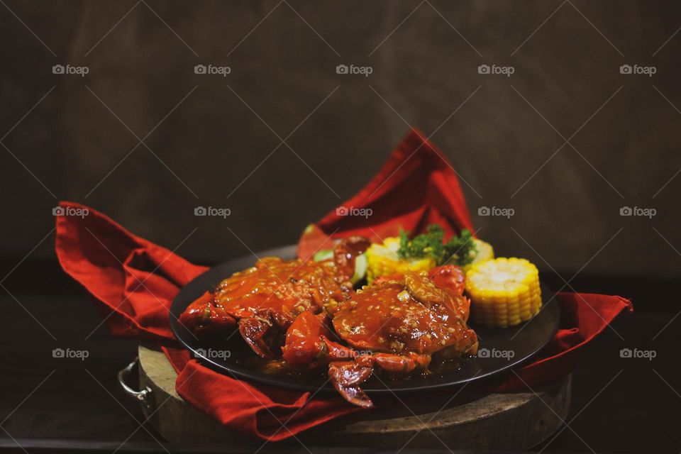 Crab flavored in creamy garlic sauce served with corn, tomatoes, lemon wedges and cherries on a black plate set on a red cloth set in a wooden setting. Asian flavor crab.