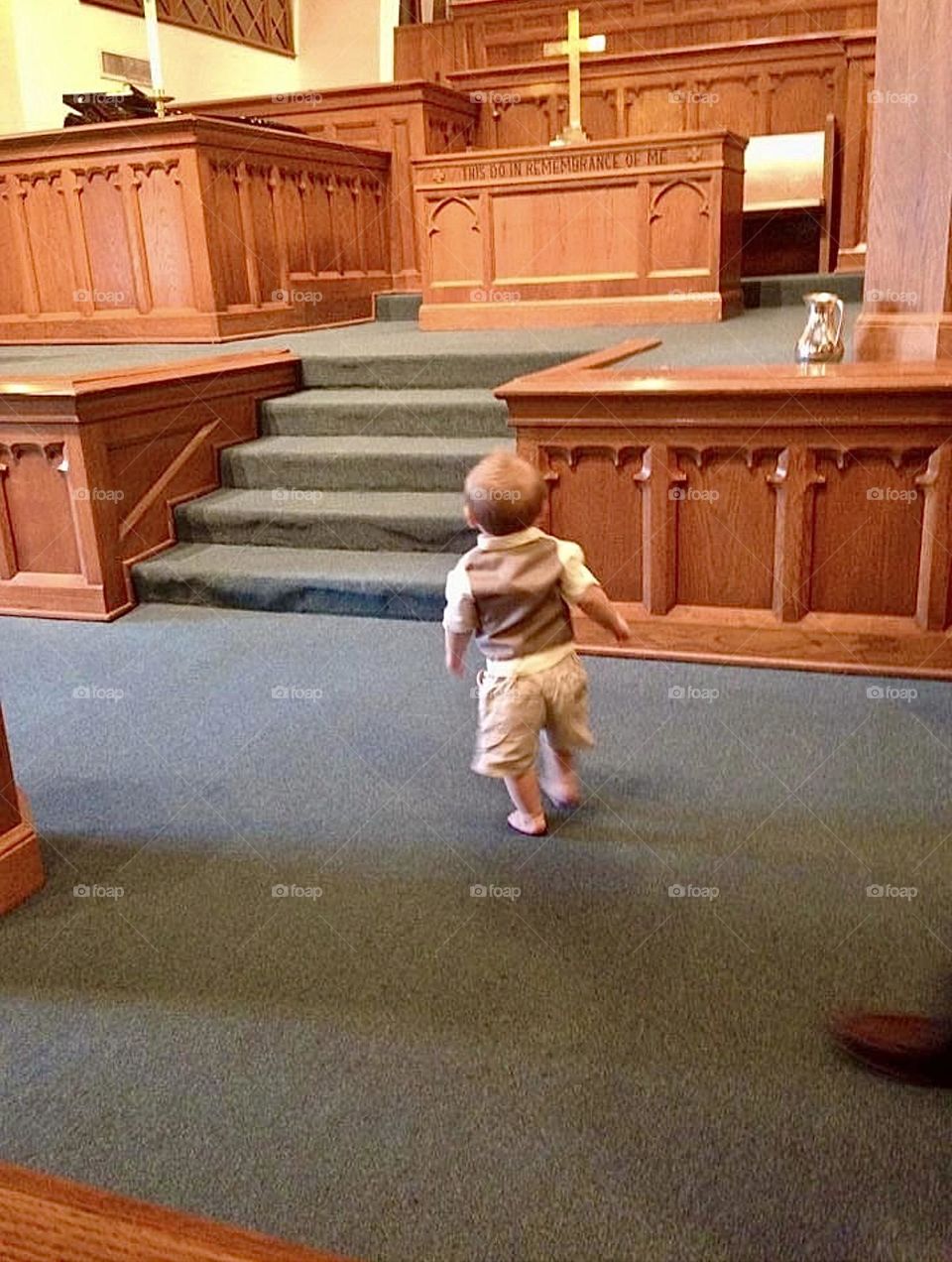 A curious toddler boy looks up to the pulpit and stage of a church. There is carpet flooring and wooden accented walls and furniture. 