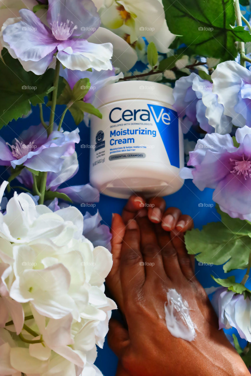 Woman applying Cerave Moisturizing Cream on blue background amongst flowers. 