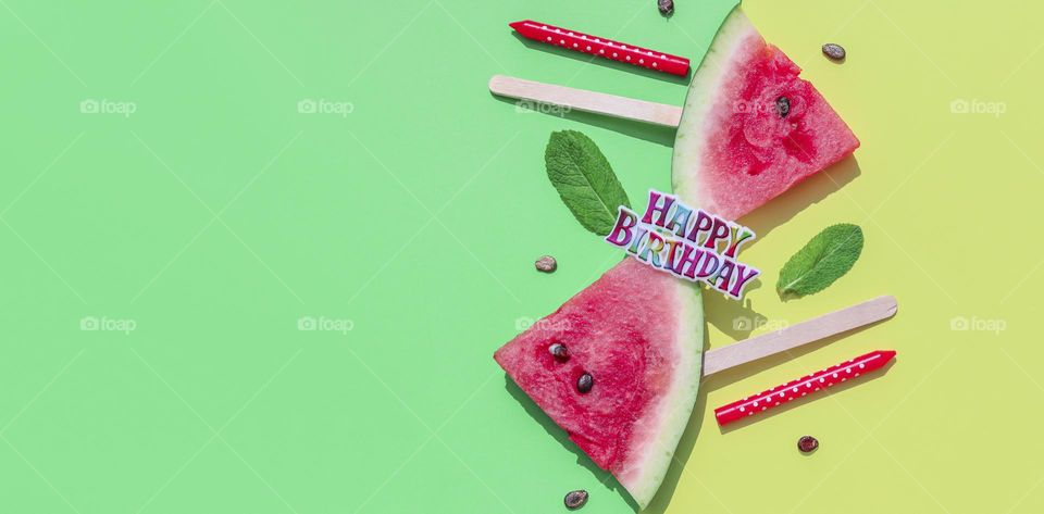 Two appetizing ripe slice of triangular shaped watermelon with mint leaves and birthday candles lie on the right on a green yellow ion with copy space on the left,flat lay close-up.Summer treats concept, popsicles, holiday banner.