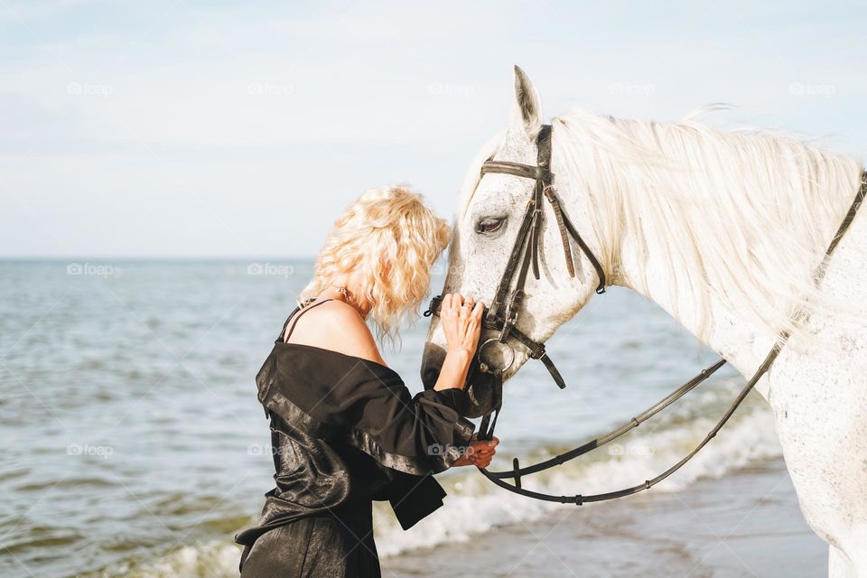 Young blonde woman in black clothes riding white horse on seascape background