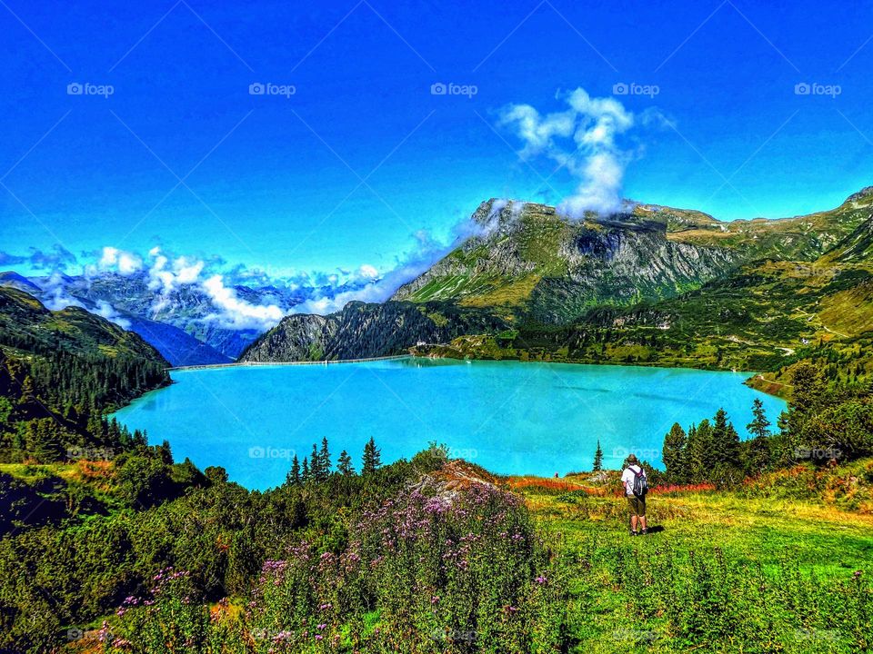 Austrian mountain landscape showing a turquoise lake surrounded by mountains, with a backdrop of bright blue sky and white fluffy clouds with colourful flowers and a hiker admiring the view in the foreground