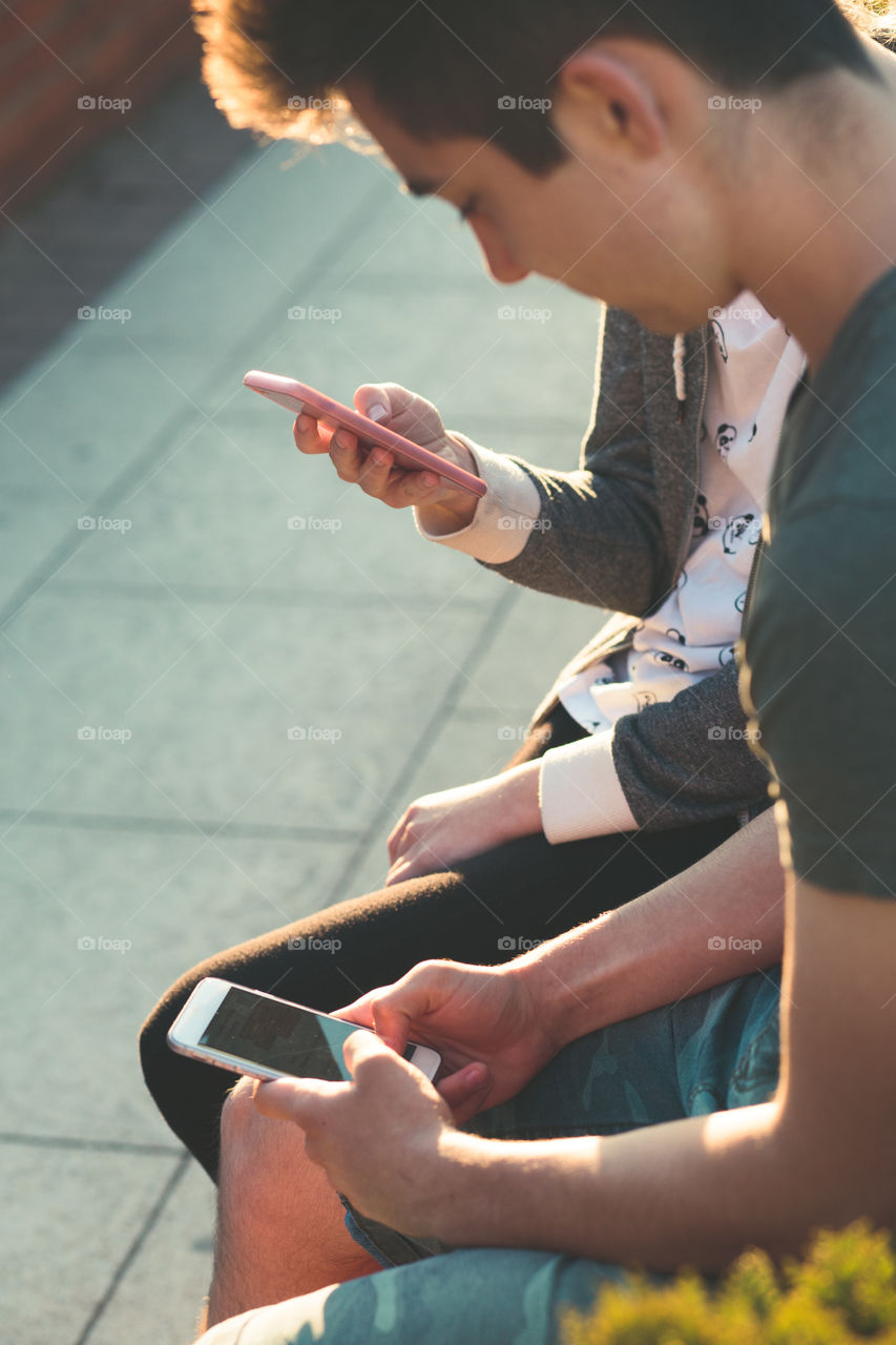 Couple of friends, teenage girl and boy, having fun together with smartphones, sitting in center of town, spending time together. Real people, authentic situations