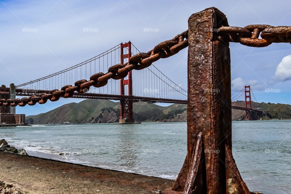 Ground level view of the Golden Gate Bridge from Fort Point in San Francisco California through the rusted iron chain link guardrails and posts that have oxidized over time appearing almost wood grain like.