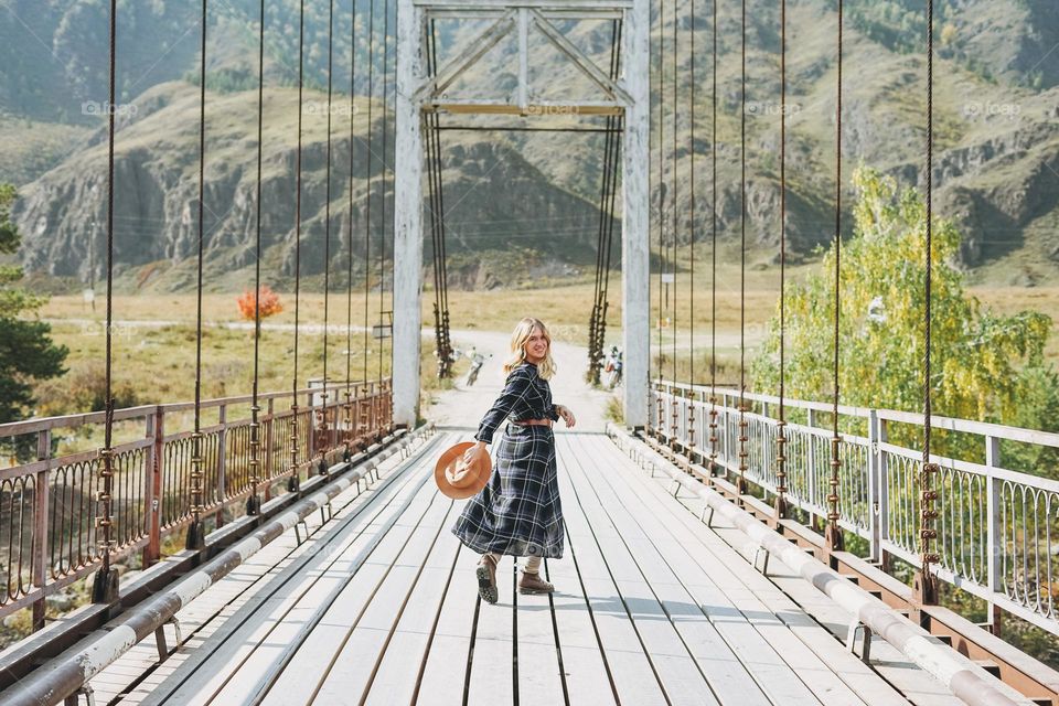Happy young blonde woman in plaid dress on bridge on river In mountain 