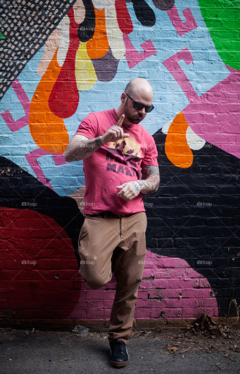 Handsome guy wearing a Hakuna Matata shirt standing against a brightly painted brick wall with sunglasses on. 