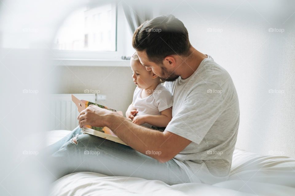 Happy father young man and baby girl little daughter having fun reading a book in children room at home