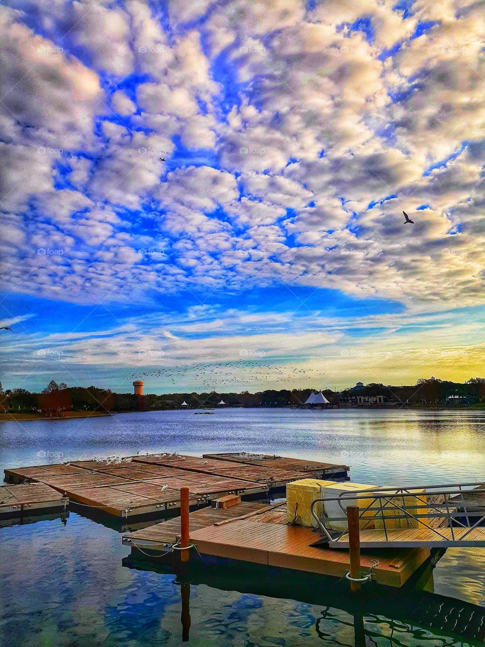 A landscape/lakescape view of the lake and dock under a beautiful blue and cloudy sky at Cranes Roost Park in Altamonte Springs, Florida.