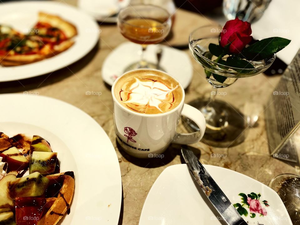 A beautiful rose latte, surrounded by plates. To the right is a rose in a beautiful cup. 