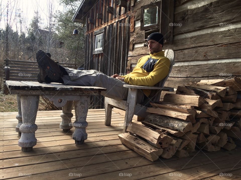 Man sitting on chair outside of log cabin next to firewood.