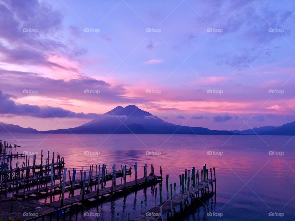 The sun gently rises over the horizon to cast a pink glow across the lake and volcano 