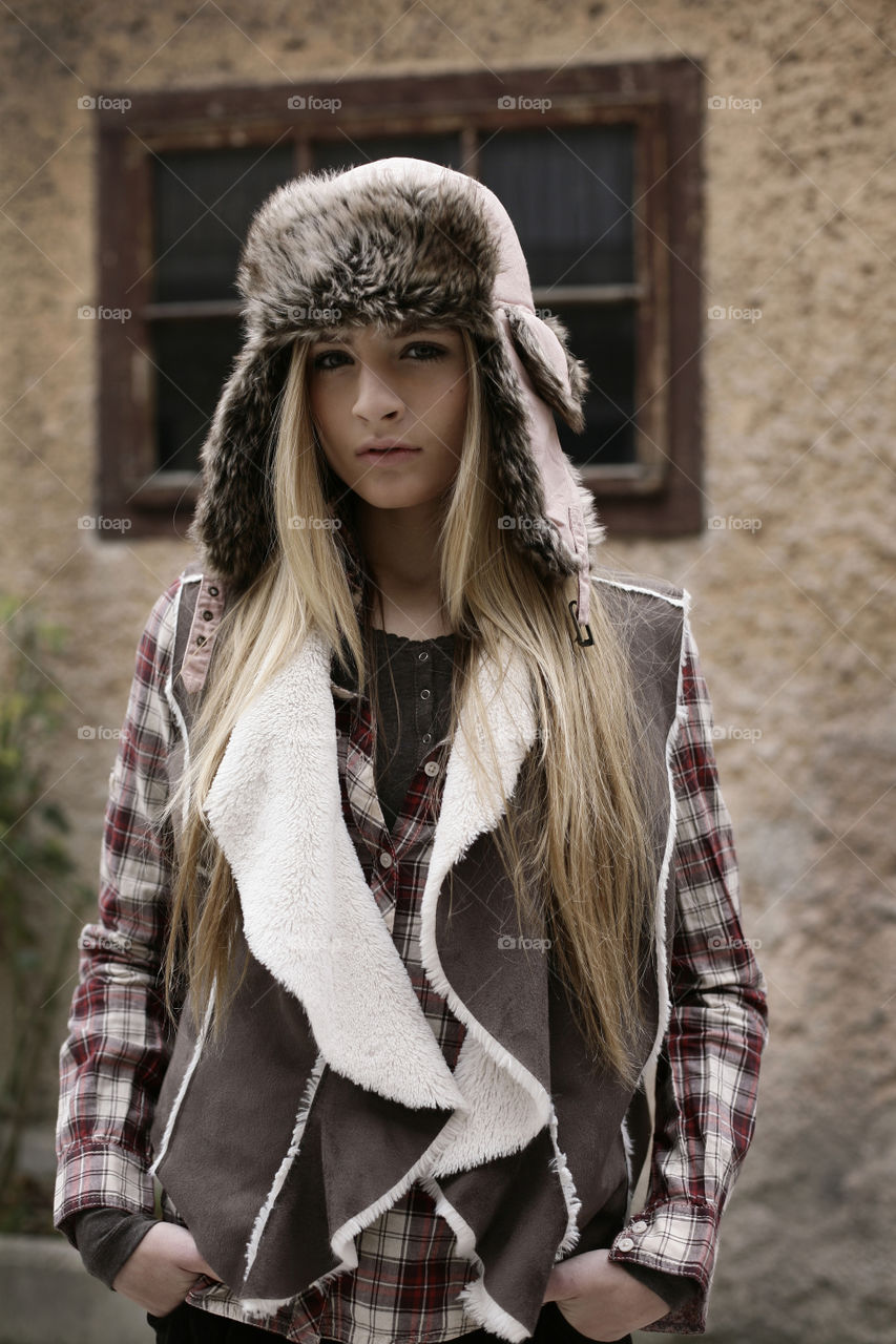 blonde girl in winter outfit. girl in front of wall and window wearing winter fur hat