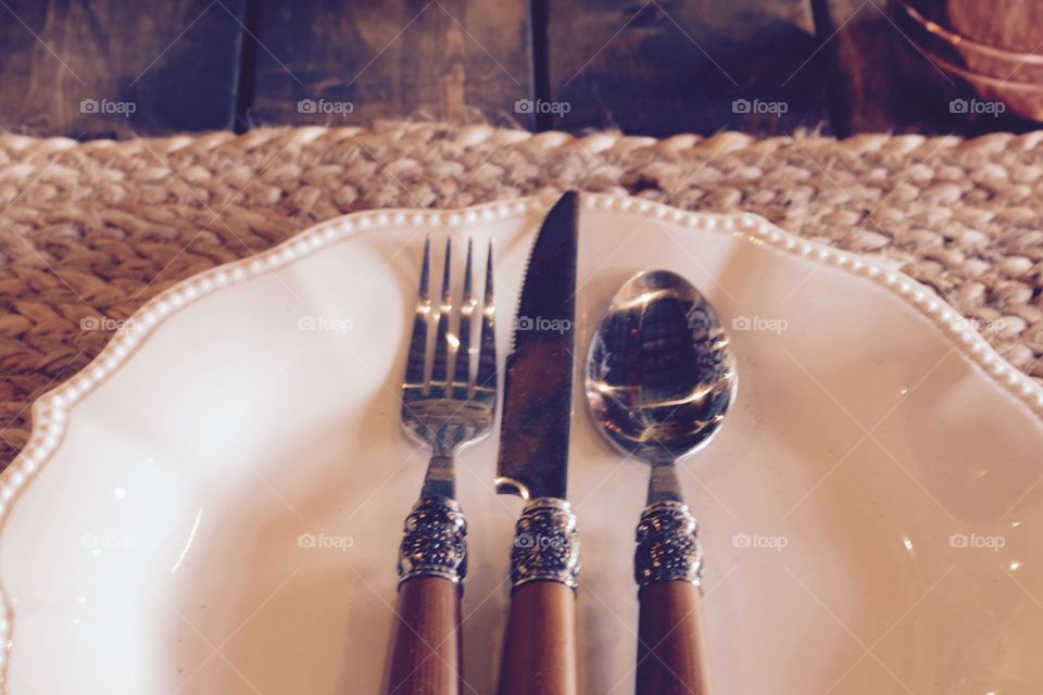 A rustic table-setting, including wooden-handled flatware trimmed with decorative stainless steel,  a scalloped, beaded white plate, and a woven placemat made of natural materials on a wooden planked table