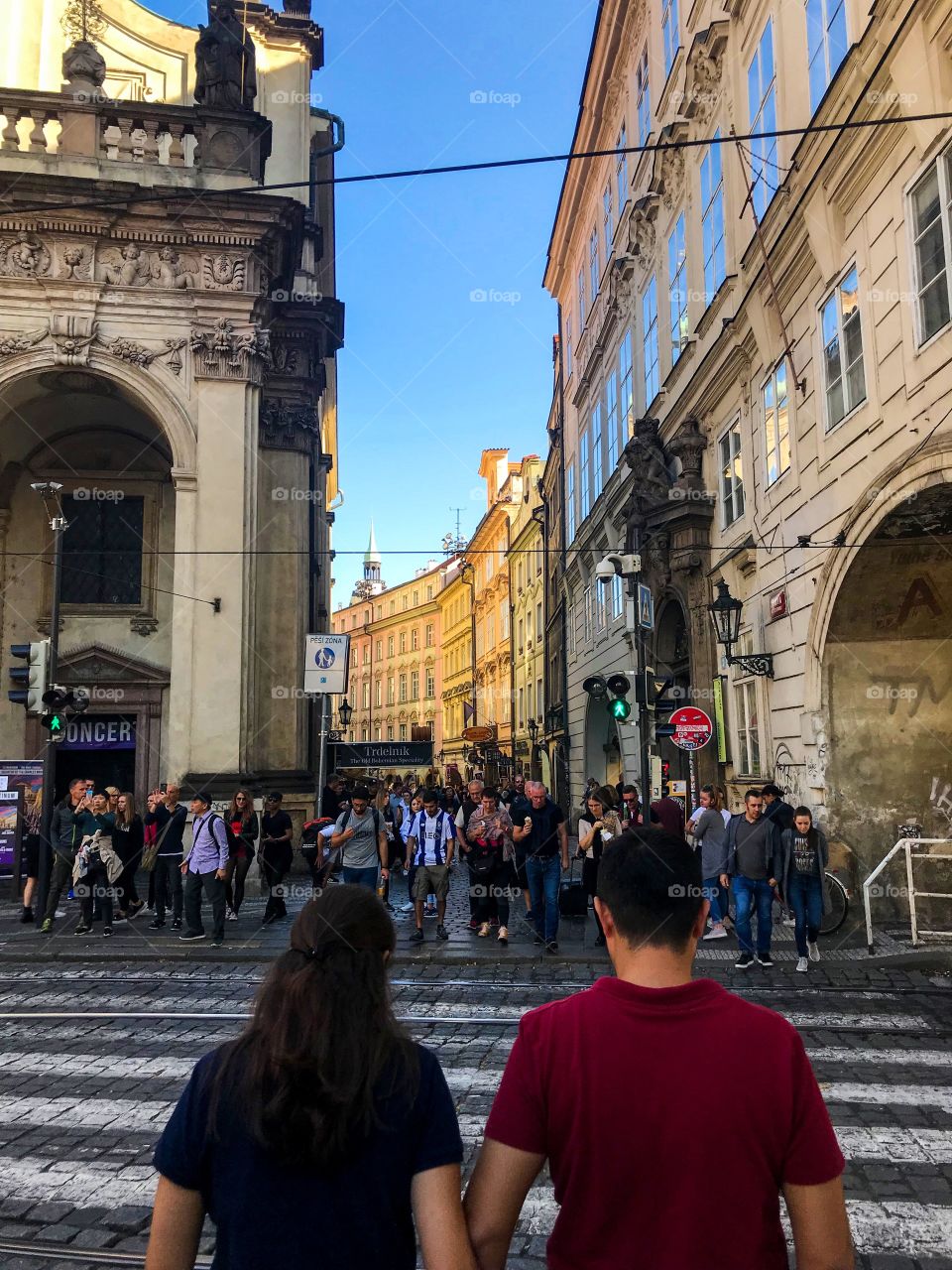 A couple is crossing the street , there are many tourists , there are many people talking, some people taking pictures . Very beautiful buildings, I am enjoying the surrounding landscape and taking pictures