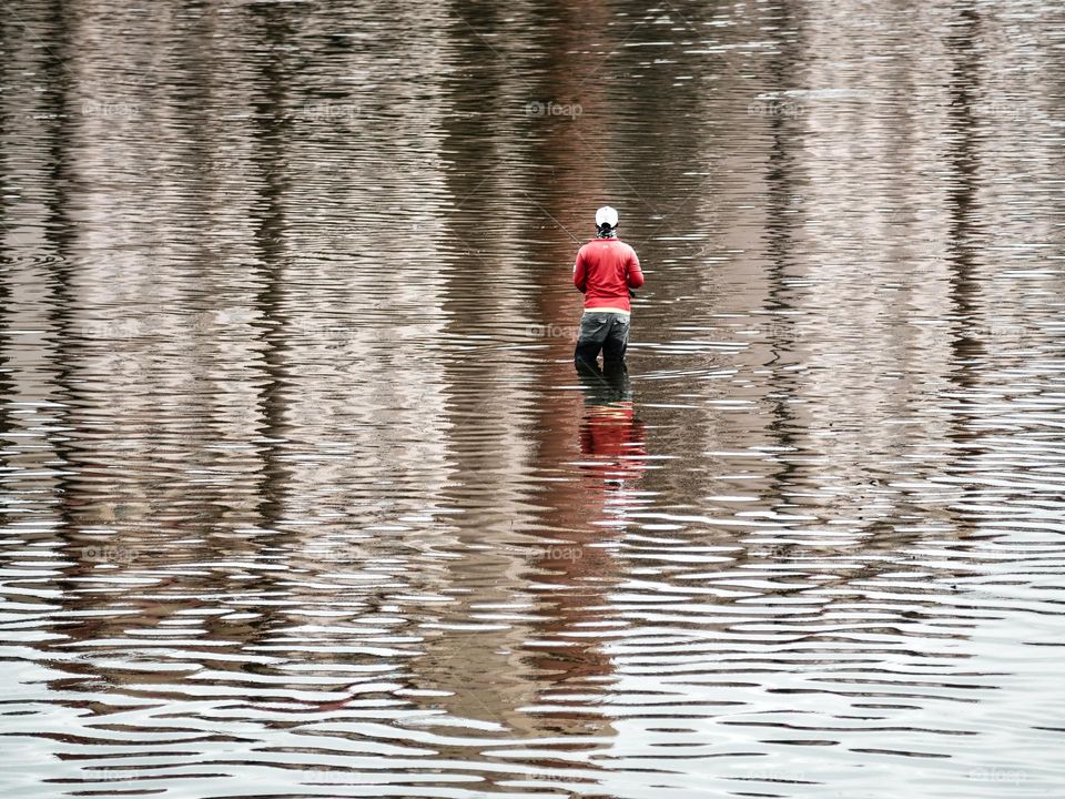 An angler fishing in the middle of a pond against building reflections on the water