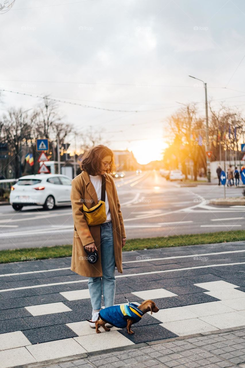 Young woman dressed with a long coat, walking her dog at sunset time in the city during winter.