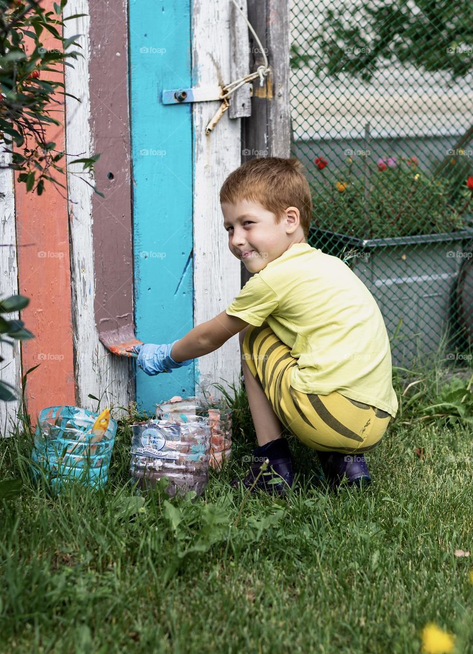 Child red-haired boy paints a wooden fence in bright colors, summer time in the countryside and holidays