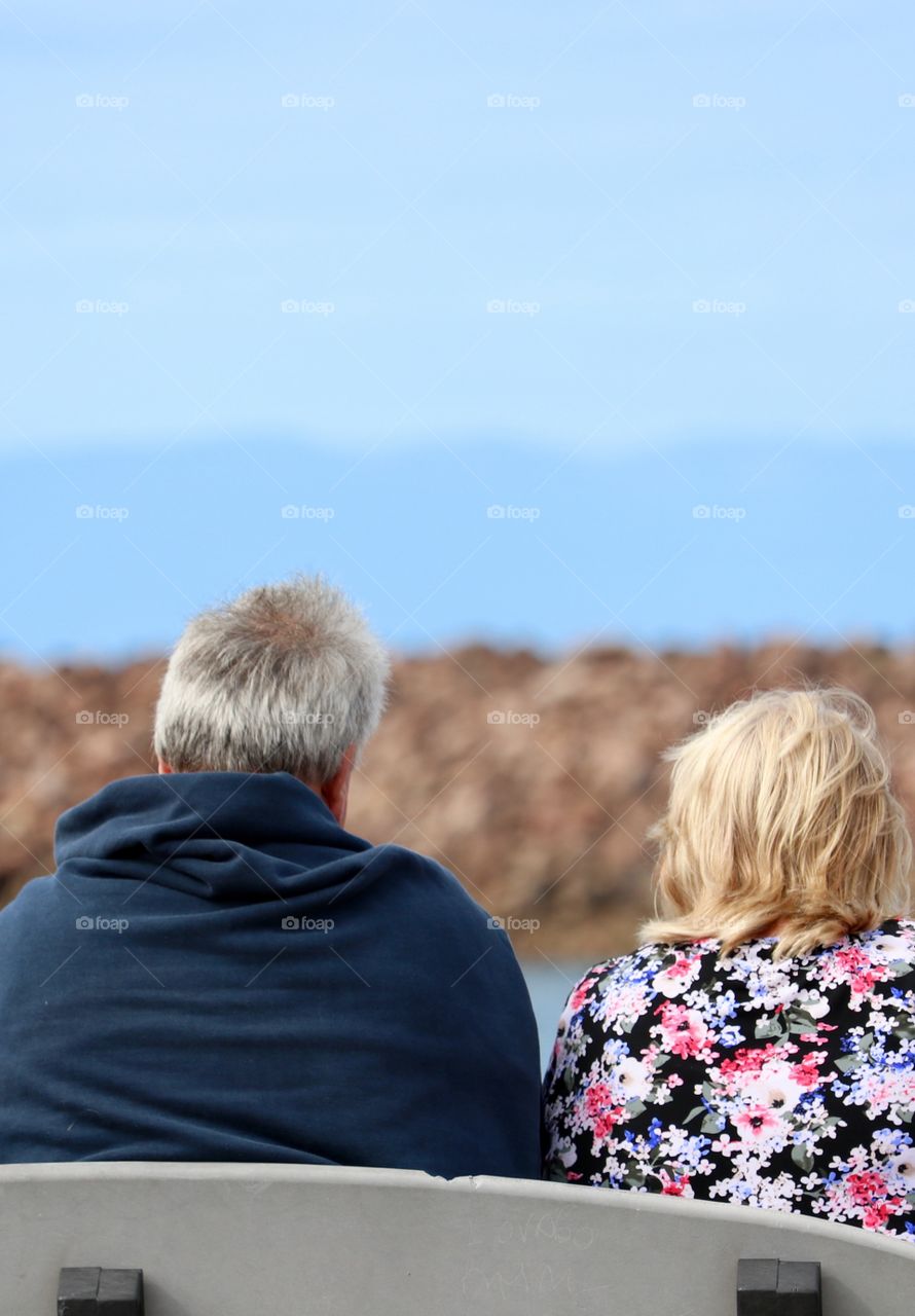 Elderly couple sitting on bench outdoors, by ocean Jetty on sky blue day 