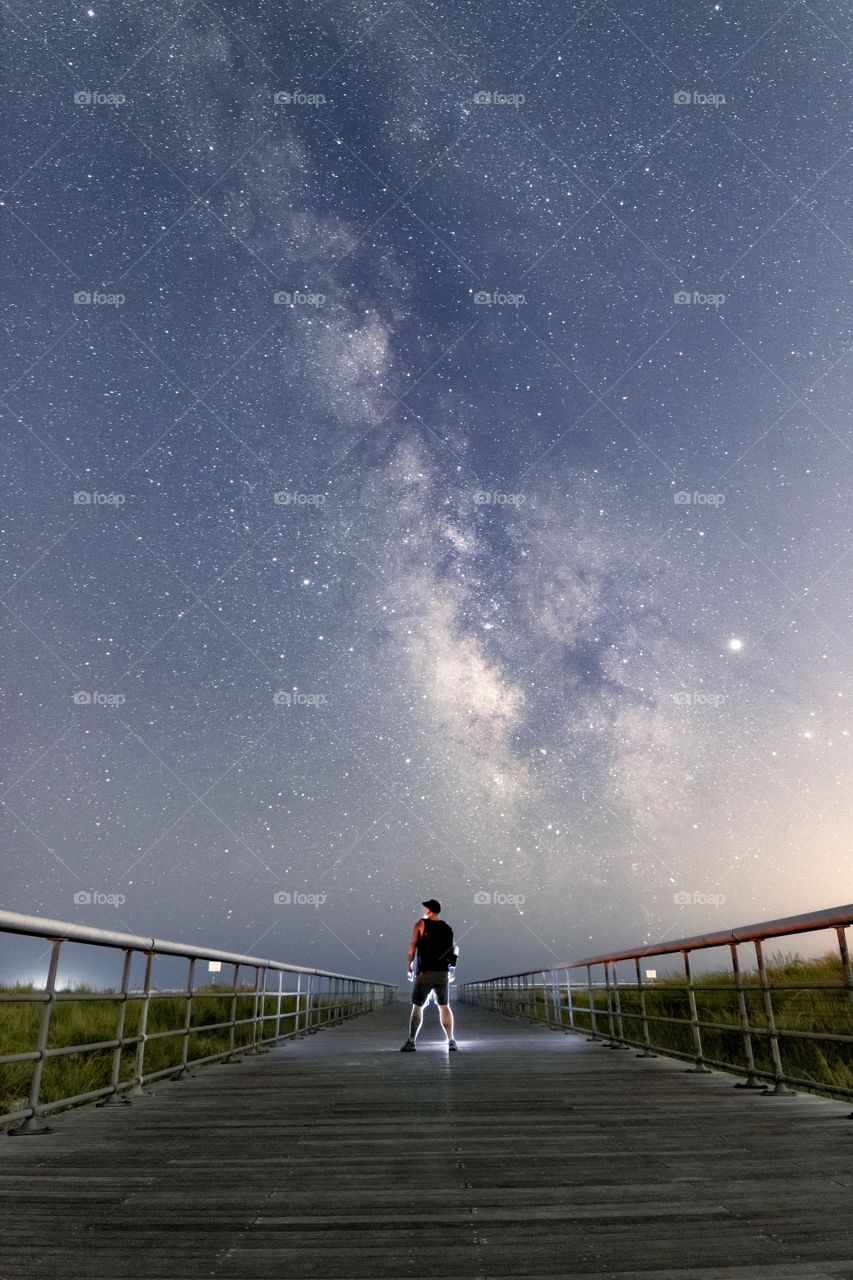 Silhouette of a man standing on a long boardwalk, leading out into the vast night sky woth the Milky Way brightly shining overhead. 
