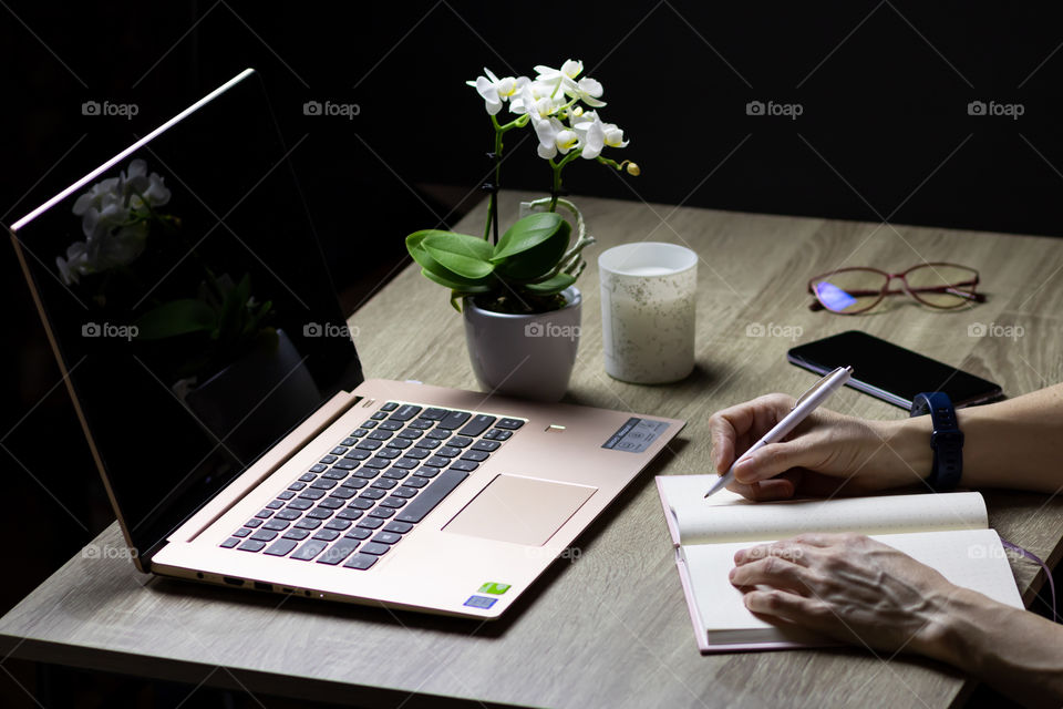 Hands are writing in a notebook next to the computer on the desktop.