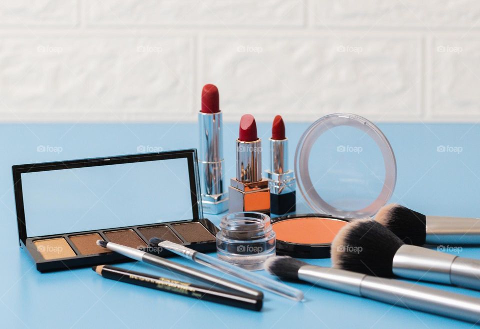 A cosmetics set of a pallet of nude eyeshadows, a round face powder, red lipsticks, soft black eyebrow shadows, makeup brushes and eye pencils lie on a blue background with a blurry white stone wall, side view. Concept female cosmetics, beauty salon.