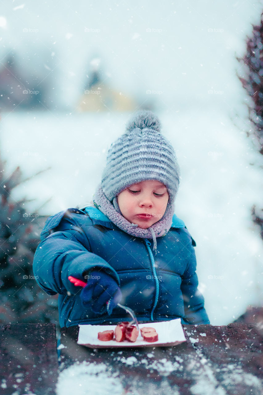 Little boy eating roasted sausage during family picnic outdoors in winter, sitting by a wooden table, wearing warm clothes, scarf and cap