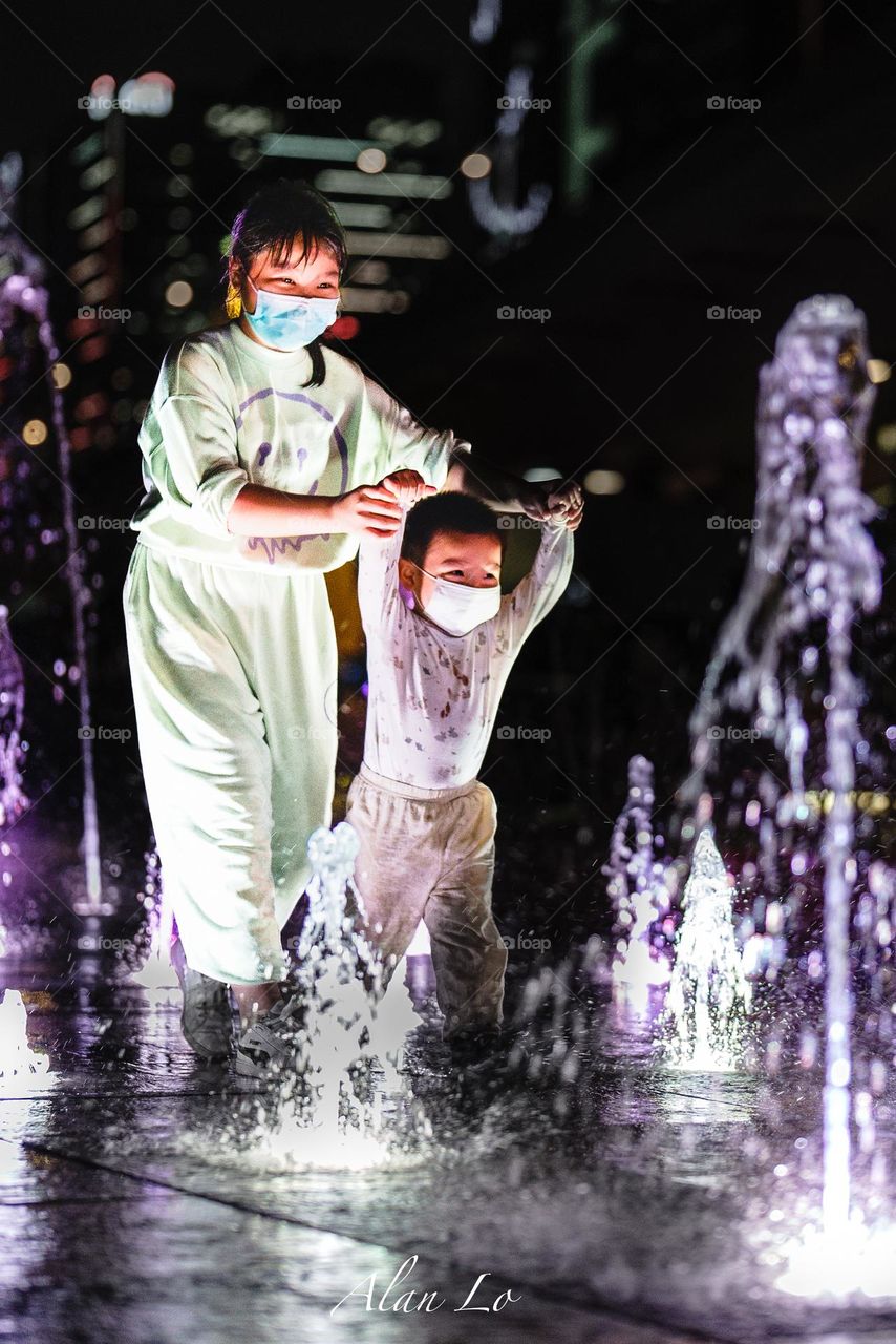 Mother’s love did not change even in the pandemic: A child delights in playing with water as a watchful adult ensures safety, both adorned with face masks to add a unique touch to this enchanting nighttime adventure.