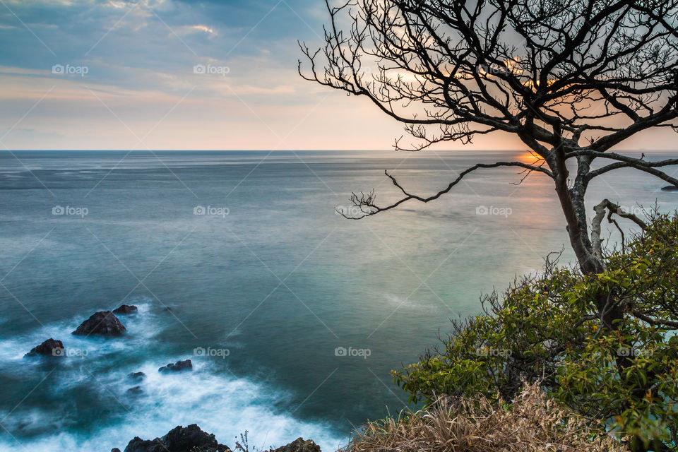 Sun setting over the Pacific Ocean near Jacó, Costa Rica with waves crashing on the rocks below and a loan tree on the cliff