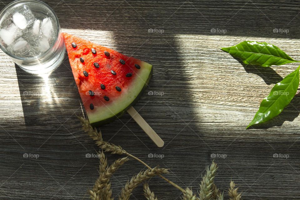 A piece of ripe, red watermelon and cold juice, a cocktail with ice and ears of wheat stand on a wooden table. Summer drinks and snacks.