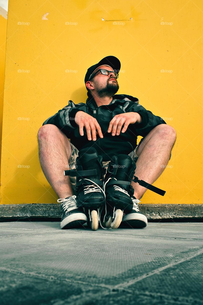 Portrait shot of bearded man with his rollerblades sitting in front of yellow wall