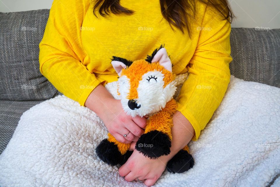 A young girl in a yellow T-shirt is sitting on a sofa, holding a cut plush fox toy and is covered with a soft blanket. Well-being in the living room. Home comfort in winter.
