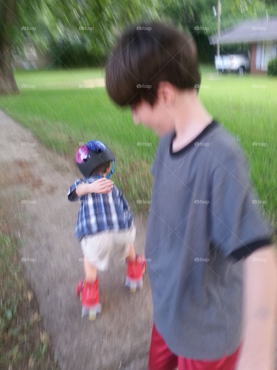 Very young boy roller skating on the sidewalk with helmet while his older brother looks on.
