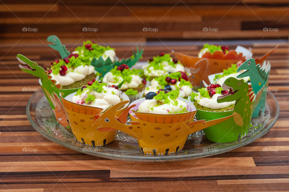 Delicious home baked dinosaur cupcakes with a fresh cream and fruits on the top served on crystal plate.