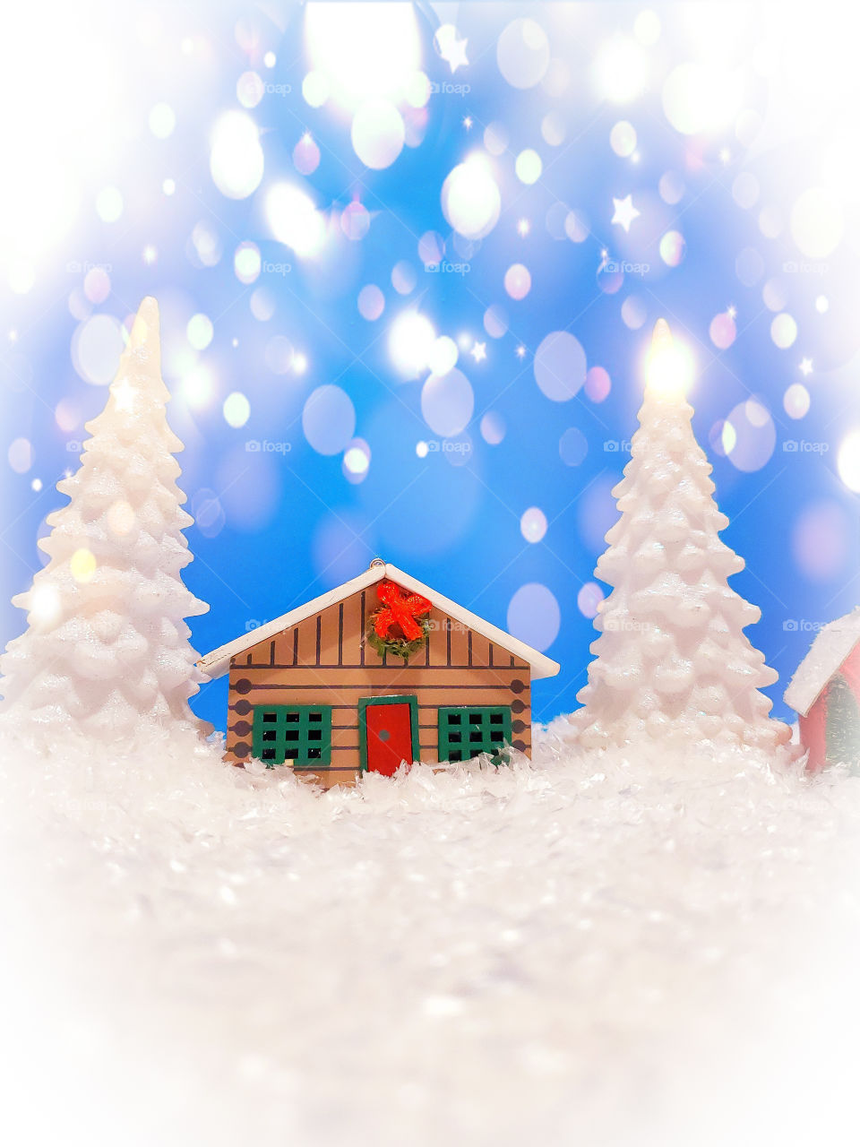 Winter miniature scene of a log cabin and two snow covered pine trees surrounded by snow and with a blue Sky illuminated by bright sparkling bokeh.