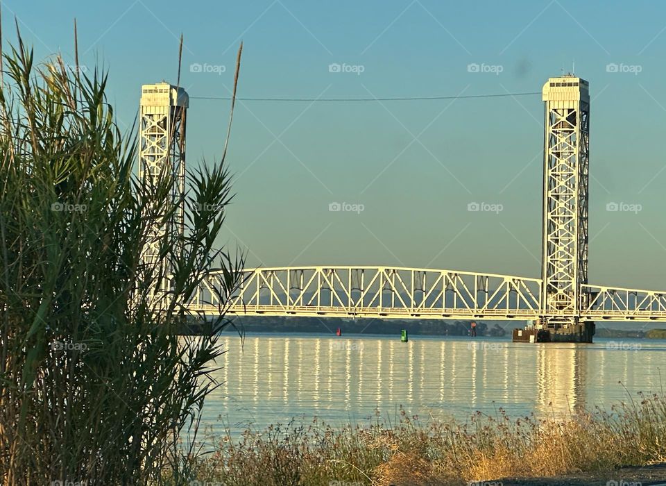 The morning sun paints the bridge with golden light, its reflection shimmering on the glassy surface of the calm river. Not a cloud disturbs the breathtaking blue sky.