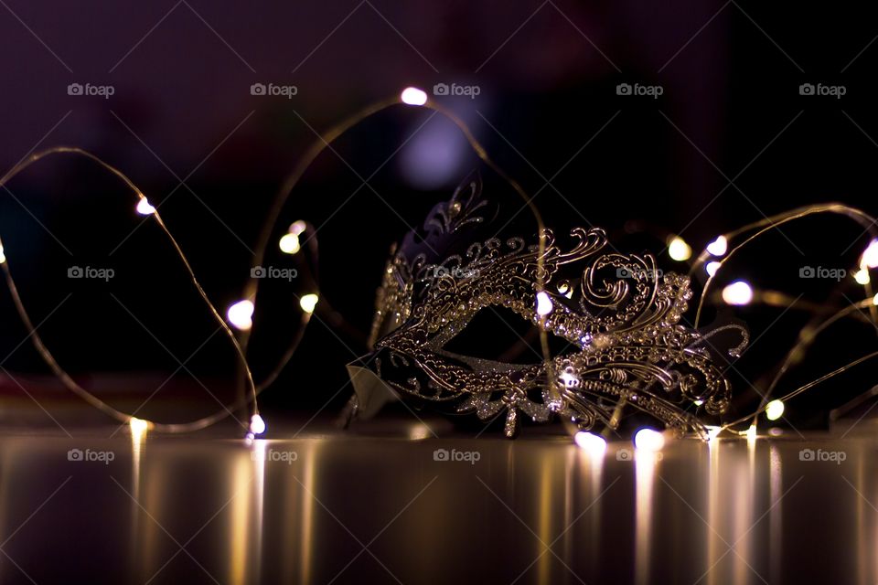 A scenic portrait of a mask lying on a table surrounded by fairy lights. the dark environment casts some mystery over the mask. it is ready to hide someones identity on a masquerade ball.
