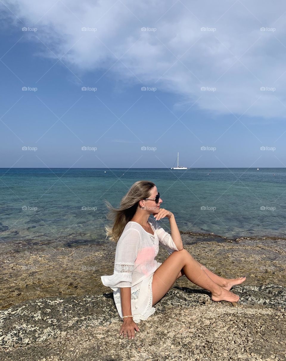 Beautiful seascape clear blue sky over the sea, a small yacht and a girl sitting on the rocks in a white dress