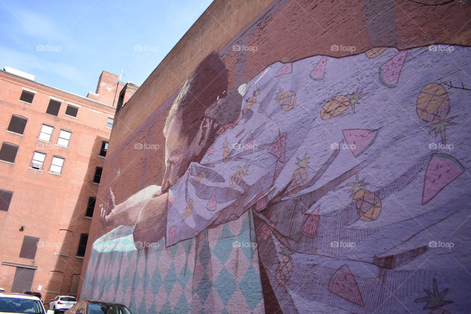 mural on brick building wall. man leaning on table with pink purple shirt with watermelon and pineapples,  rat on shoulder
