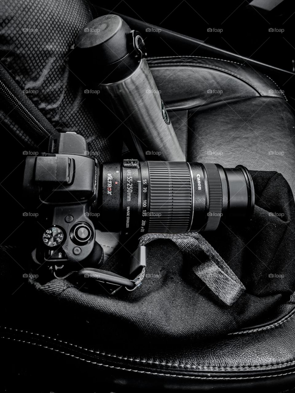 Photo hunting. A black camera with a dark silver tumbler on a black car seat