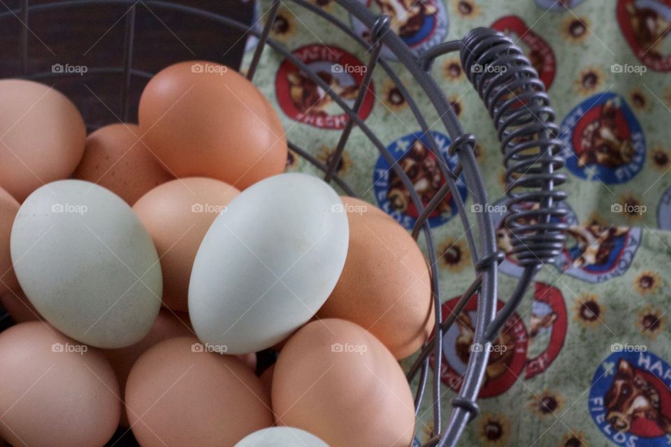 Overhead view of farm-fresh blue and brown eggs in a wire basket and dairy-themed apron with pocket on a dark wooden surface