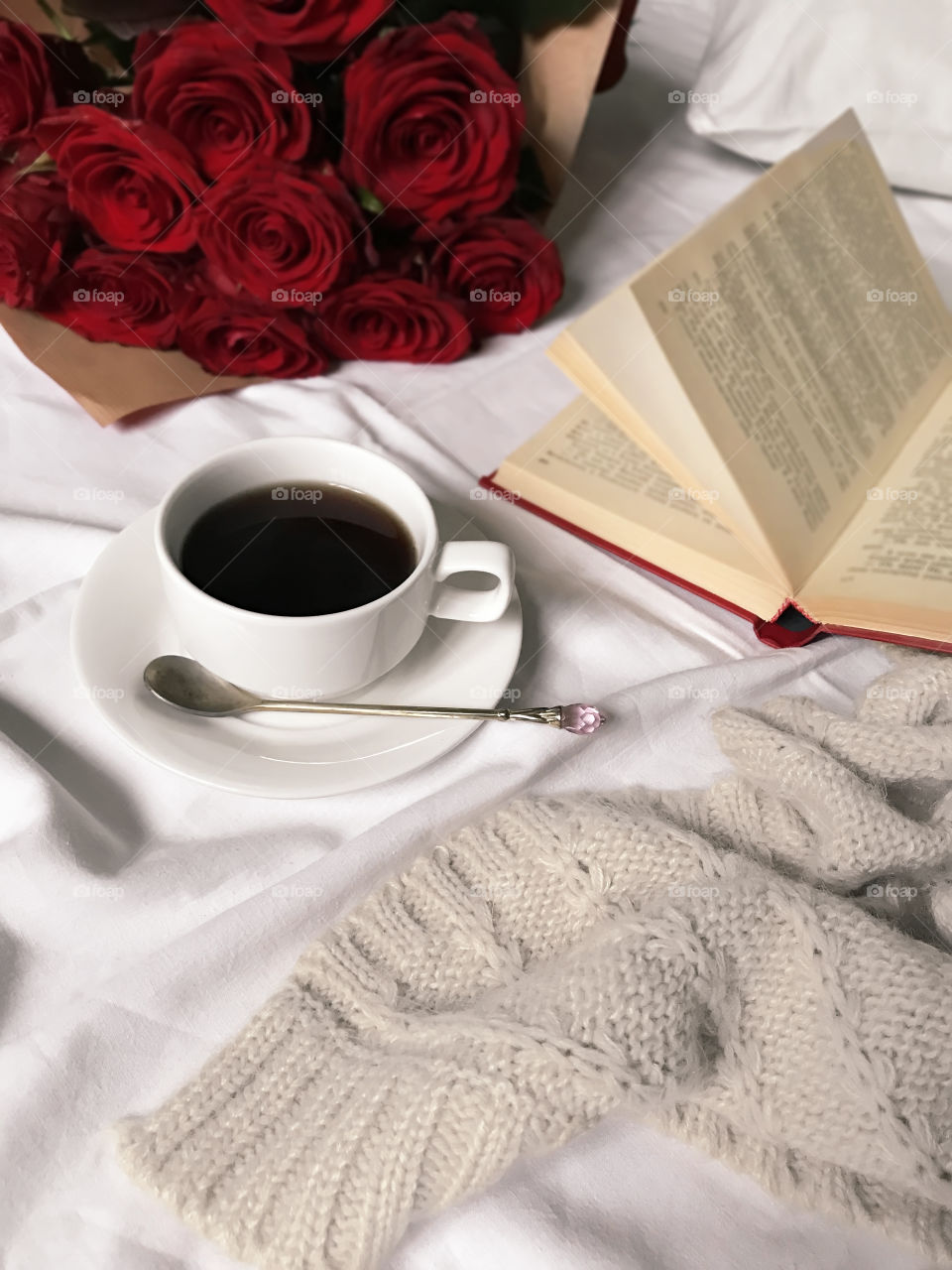 White cup of coffee with red roses and red book in cozy white bed 