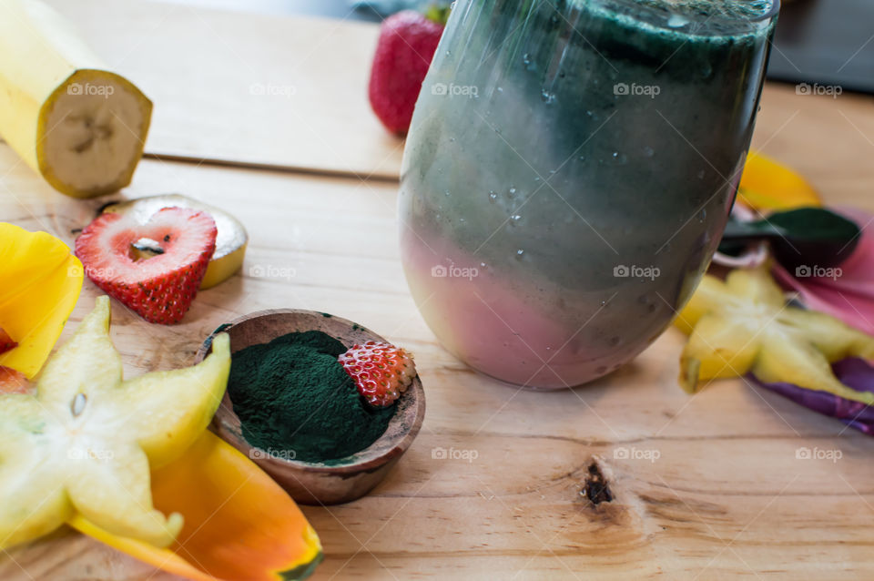 Rainbow dream smoothie with pink, yellow and green with bowl of protein powder next to glass on wood background surrounded by fresh fruit: strawberry, banana, star fruit and pastel Colors 