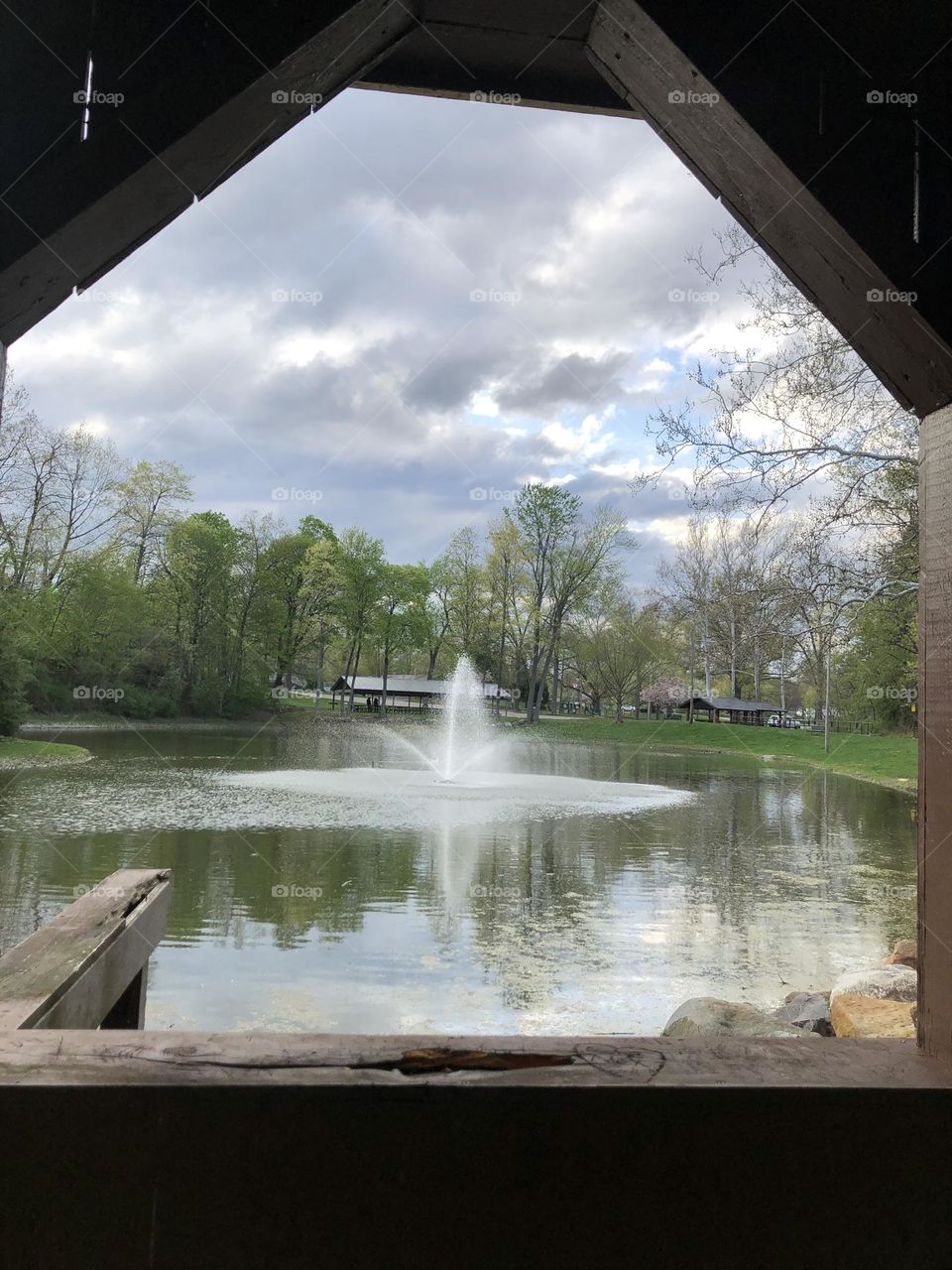 A look through the covered bridge, the fountain flowing freely into the pond which has the reflection of the sky in the rippling in pond waters 